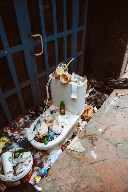 A white ceramic toilet with a closed lid and a rectangular cistern, positioned outdoors against a dark, metal fence with vertical slats and a curved handle on the left side. The toilet is cluttered with various waste items, including an empty plastic cup, a wine glass, and a cardboard tube, all resting on and around the cistern. The surrounding area is littered with used paper, plastic bottles, cans, and food wrappers, scattered on the concrete ground and partially blocking access. A small green glass bottle is placed on the toilet bowl, amidst the rubbish. To the right, the surface transitions from the rough, cracked concrete pavement to a slightly raised section, with additional waste and a black plastic bag partially out of frame. The overall scene exemplifies a site where private waste clearance services, such as those provided by Waste Clearance Ilford, might be employed to address environmental or access issues caused by unmanaged refuse accumulation.