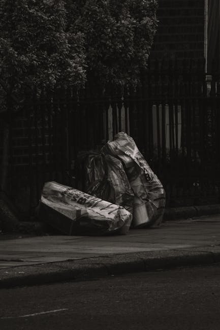 A group of black plastic trash bags filled with waste leaning against a black metal fence along a sidewalk. The bags appear to contain mixed rubbish, with some showing partially visible printed labels and creased, crumpled textures. The bags are positioned close together, with the larger ones slightly leaning and appearing somewhat heavy. In the background, there are leafy trees that cast shadows, and part of a building with a dark exterior is visible behind the fence. The scene is outdoors on a paved street or pavement area, indicating an area where waste collection or disposal might take place, potentially managed by private waste removal services such as Waste Clearance Ilford. The overall lighting is subdued, giving a muted tone to the image, which emphasizes the waste bags and surrounding environment.