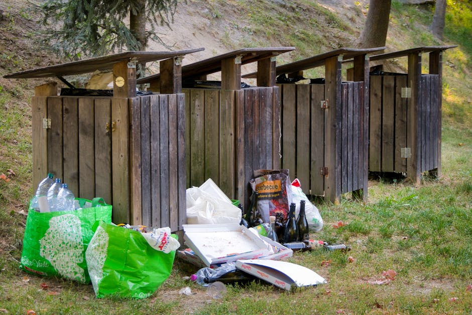 A row of four wooden outdoor waste bins with slanted roofs are positioned on a grassy area near a wooded slope, with trees and greenery in the background. The bins, constructed from vertical wooden planks with visible hinges and small locks, appear weathered and darkened by exposure to the elements. Several of the bins are filled with trash, with some waste spilling onto the ground in front. Among the discarded items are empty plastic bottles, a partially opened cardboard pizza box, a crumpled paper bag, and other miscellaneous litter including glass jars and plastic packaging. The trash is loosely arranged, with some items leaning against the bins and others lying directly on the grass, which is speckled with fallen leaves. The scene conveys a sense of informal waste disposal, possibly an alternative to official rubbish collection, set in a natural outdoor environment illuminated by soft daylight. Waste Clearance Ilford’s name is subtly referenced through the context of visible rubbish removal services, highlighting private or on-site clearance practices for outdoor waste management.