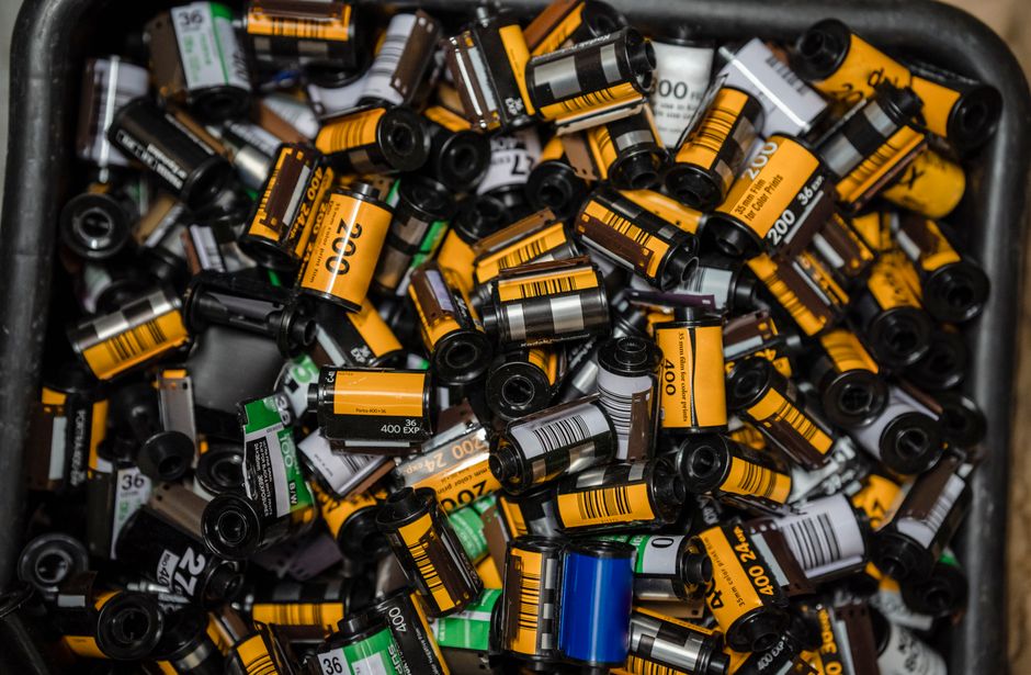 A close-up view of a large black plastic container filled with numerous used photographic film canisters and developer bottles. The cylindrical objects vary in size and primarily feature yellow, black, white, and green labels with barcodes and text indicating film numbers and expiry dates. Some cans have metallic silver caps, while others have black plastic lids. The canisters are tightly packed, overlapping each other, with some leaning against the edges of the container. The environment suggests an indoor setting, possibly a storage area or waste collection site, with no visible background beyond the container's edge. The lighting is even, highlighting the glossy textures of the plastic and metallic components, emphasizing the disorganized accumulation typical of waste requiring collection or disposal. Waste Clearance Ilford often manages such bulky or specialty waste items, including chemical containers from photographic or similar industries, as part of their rubbish removal services, supporting private disposal or alternative waste handling options.