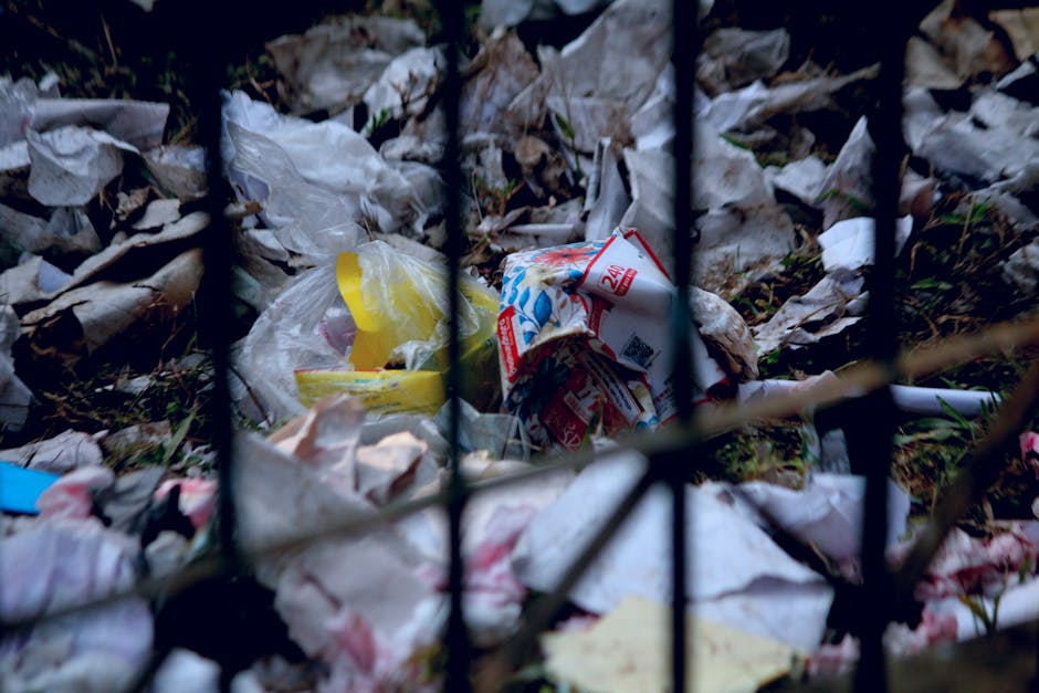 A close-up view of a pile of scattered waste materials behind a black metal fence, including crumpled plastic bags, torn paper, and discarded packaging, with some items partially buried under dried leaves and soil. The waste appears to be a mix of household rubbish, such as food wrappers, paper cartons, and plastic packaging, all exhibiting varying degrees of crumpling, tearing, and weathering. The background shows an outdoor environment with more debris scattered on the ground, suggesting an unmanaged or illegal dumping site. The lighting is natural, with soft shadows indicating daylight, and the scene emphasizes the importance of proper rubbish disposal and waste management, possibly related to private waste collection services like Waste Clearance Ilford, who handle rubbish removal beyond standard council collection methods.