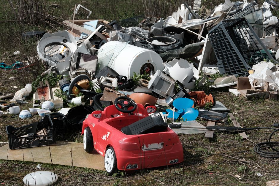 A large pile of mixed household waste and discarded items situated outdoors on soil and grass, including a weathered red plastic children's toy car in the foreground with visible scratches and chipped paint, surrounded by various broken appliances, electronic devices, and household objects such as a white washing machine drum, plastic buckets, and black plastic crates. The background features an assortment of white, black, and beige household items, including disassembled furniture parts, a microwave, and appliances, all arranged chaotically in a heap. The scene is set in an outdoor environment with sparse vegetation and natural sunlight illuminating the debris, highlighting the textures of the worn and damaged materials. This clutter of non-recyclable household waste exemplifies a scenario often addressed by private waste clearance services like Waste Clearance Ilford, which provides alternatives to local authority rubbish removal for reducing illegal dumping and managing bulk rubbish effectively.