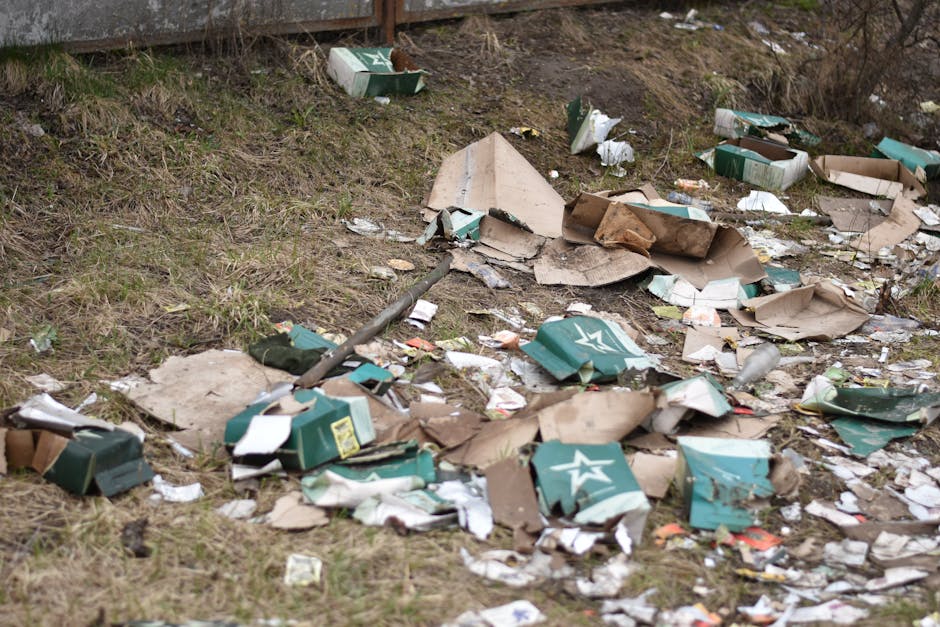 A patch of ground adjacent to a wooden fence shows scattered rubbish including torn and crushed cardboard boxes with green and white branding, some flattened and others partially collapsed, along with various paper and plastic debris. The boxes are irregularly strewn across on a mix of sparse grass and bare soil, with evidence of weathering and dirt on their surfaces. Among the debris, there are small pieces of paper and plastic wrappers, with a few partially buried in the ground. A long, dark metal pole lies diagonally across the scene, partially covered by the rubbish. The environment appears outdoors, with natural daylight illuminating the clutter, which is indicative of informal disposal or unmanaged waste accumulation, often associated with private rubbish clearance or illegal dumping. The scene suggests a need for professional rubbish removal services, such as those offered by Waste Clearance Ilford, to manage and clear the waste effectively and responsibly.