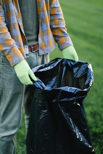 A person, wearing a plaid shirt in shades of yellow, grey, and blue over a green top, is standing outdoors on a grassy area. They are wearing light grey trousers with a brown belt and bright green gloves. The individual is holding open a black rubbish bag, made of shiny plastic material, which is partially filled and positioned upright on the ground. The surrounding environment features lush, well-maintained green grass, suggesting an outdoor setting such as a park or garden. The image conveys the act of rubbish collection or waste disposal, relevant to private waste handling services offered by Waste Clearance Ilford, emphasizing environmentally responsible rubbish removal or on-site clearance practices, with a focus on proper waste containment and disposal in an open outdoor space.