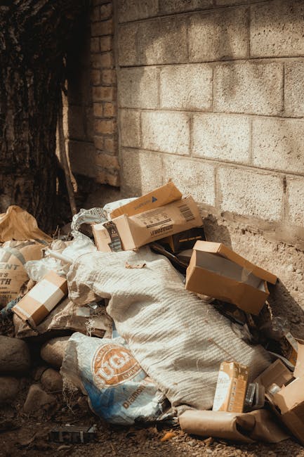 A pile of mixed cardboard boxes, some flattened and others partially opened, along with large white and beige waste sacks, all situated against a rough brick wall and a concrete block wall in an outdoor setting. The cardboard boxes vary in size and are light brown with printed labels or barcodes visible, indicating they are for disposal or recycling. The sacks appear weathered and are filled with unidentifiable waste materials. The scene is illuminated by natural daylight, casting soft shadows on the ground, which consists of gravel and small stones. This collection of rubbish suggests an instance of on-site waste clearance, possibly undertaken by Waste Clearance Ilford, highlighting the need for professional rubbish removal services focusing on private waste handling and alternative disposal methods outside local authority collection. The environment appears to be a backyard or side alley adjacent to a building, emphasizing the context of non-commercial private waste disposal or clearance services. Overall, the scene reflects a typical example of outdoor waste accumulation awaiting removal or recycling.
