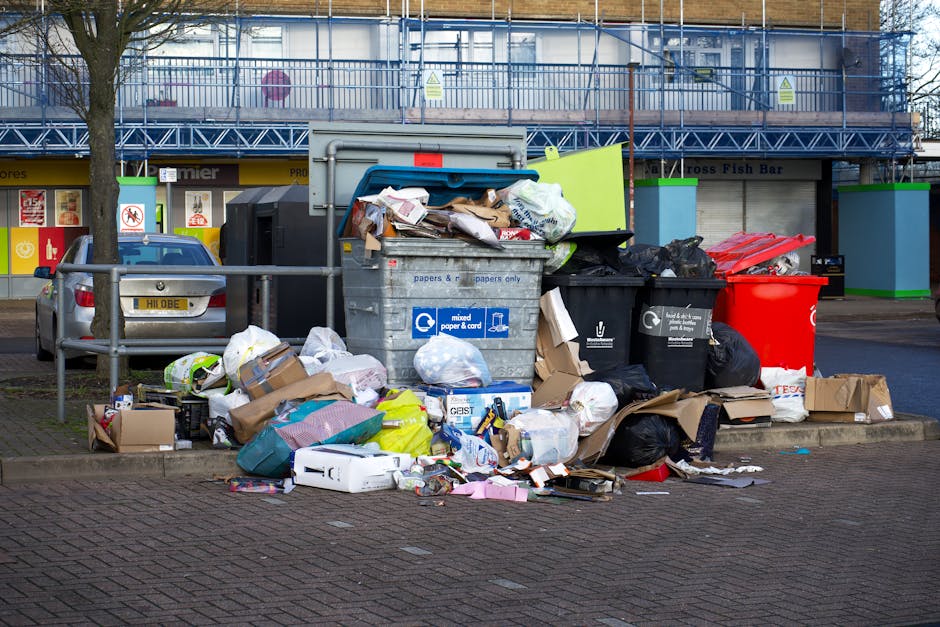 A collection of overflowing waste bins and scattered rubbish on a paved sidewalk in an urban commercial area. The scene includes multiple black, brown, and red plastic rubbish bins filled or spilling over with mixed waste, including cardboard boxes, paper, plastic bags, and packaging materials. Some items, such as a white cardboard box and crumpled paper, are strewn across the ground around the bins. The backdrop features a building with a storefront, possibly a shop or café, with signage visible behind a glass façade. A parked silver car is positioned close to the rubbish area, partly obscured by a metal railing that separates the waste from the pavement. The environment is illuminated by natural daylight, highlighting the cluttered, unkempt appearance of the waste collection point, which appears suitable for a private rubbish removal service like Waste Clearance Ilford in the context of alternative waste handling options near Ilford Station.